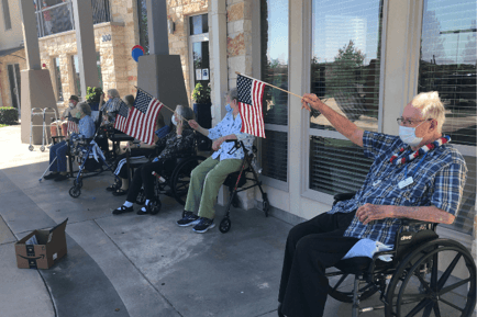 A row of seniors wearing masks and waving American flags outdoors in front of Arbor Terrace Lakeway — a festive Labor Day car parade at this beloved senior care community Lakeway TX.