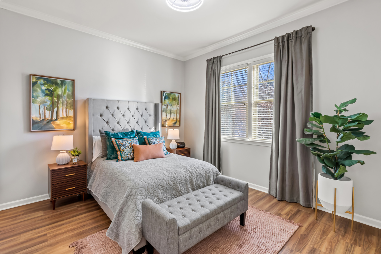 A cozy model bedroom at this senior living community in Acworth, GA, featuring a tufted headboard, warm wood floors, soft drapery, and a cheerful houseplant that give the space a homey feel.