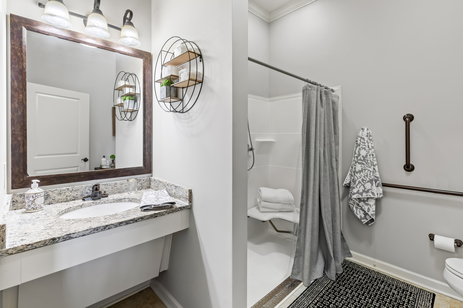 A clean and accessible model bathroom at this Acworth assisted living community, featuring a wide vanity, framed mirror, roll-in shower, and thoughtful safety features throughout.