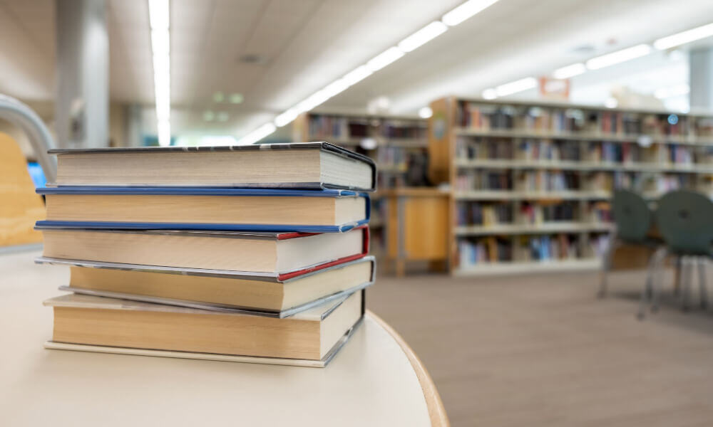 A bright and well-stocked library with neatly arranged bookshelves and a stack of books in the foreground, reflecting the enriching, engaging senior lifestyle programs available near Acworth, GA.