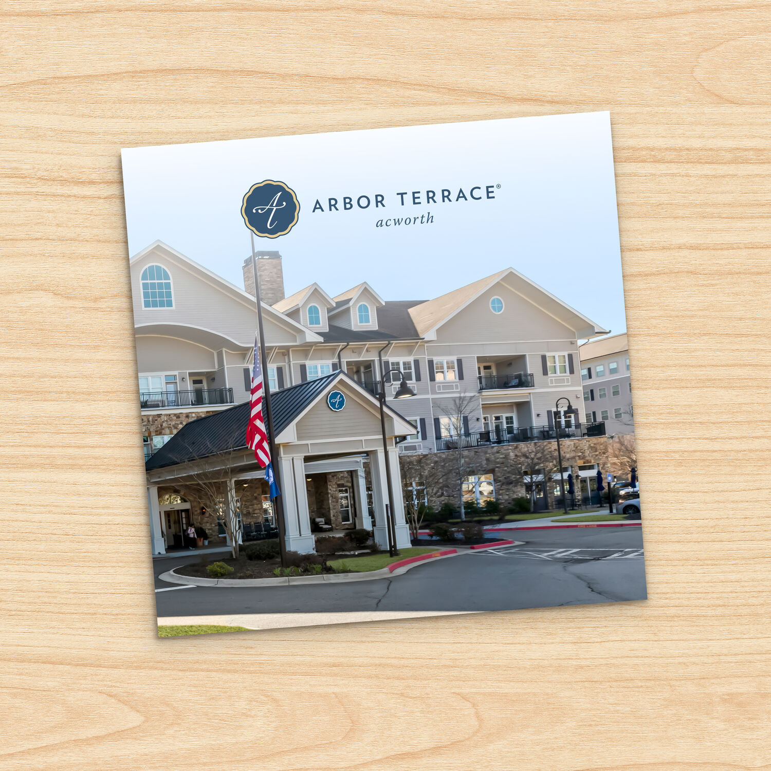 Square brochure cover for Arbor Terrace Acworth, angled on a light wood surface. The cover photo shows a large multi-story senior living building with gray siding and stone accents, a covered entrance porch, and an American flag on a pole in the circular driveway.
