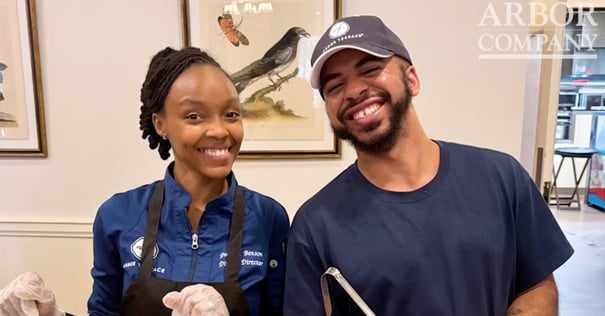 Two smiling Arbor Terrace staff members in navy uniforms pose together indoors, with a framed bird illustration on the wall behind them and the Arbor Company logo in the corner.