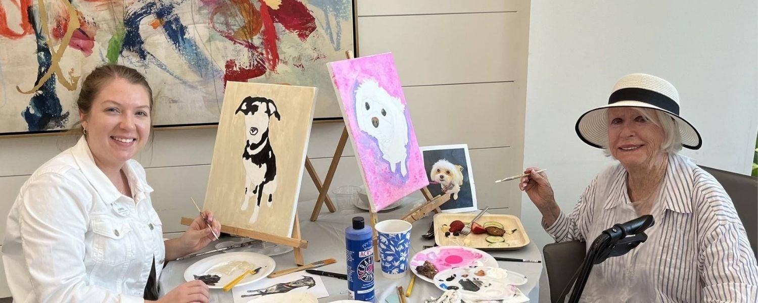 Two smiling women paint pet portraits side by side at easels, one in a white jacket working on a black and white dog, the other in a sun hat and striped shirt painting a fluffy white pup, a creative afternoon in active senior communities in Atlanta, GA and top-rated senior living in Atlanta, GA.