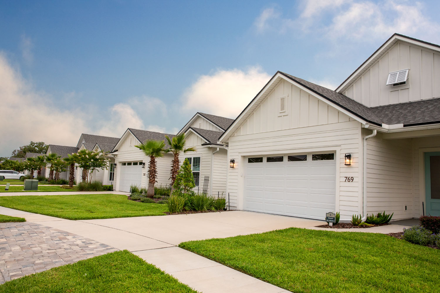 A row of charming white cottage homes with two-car garages and manicured lawns sits beneath a bright blue sky, showcasing the residential living options at The Lakeside at Amelia Island.