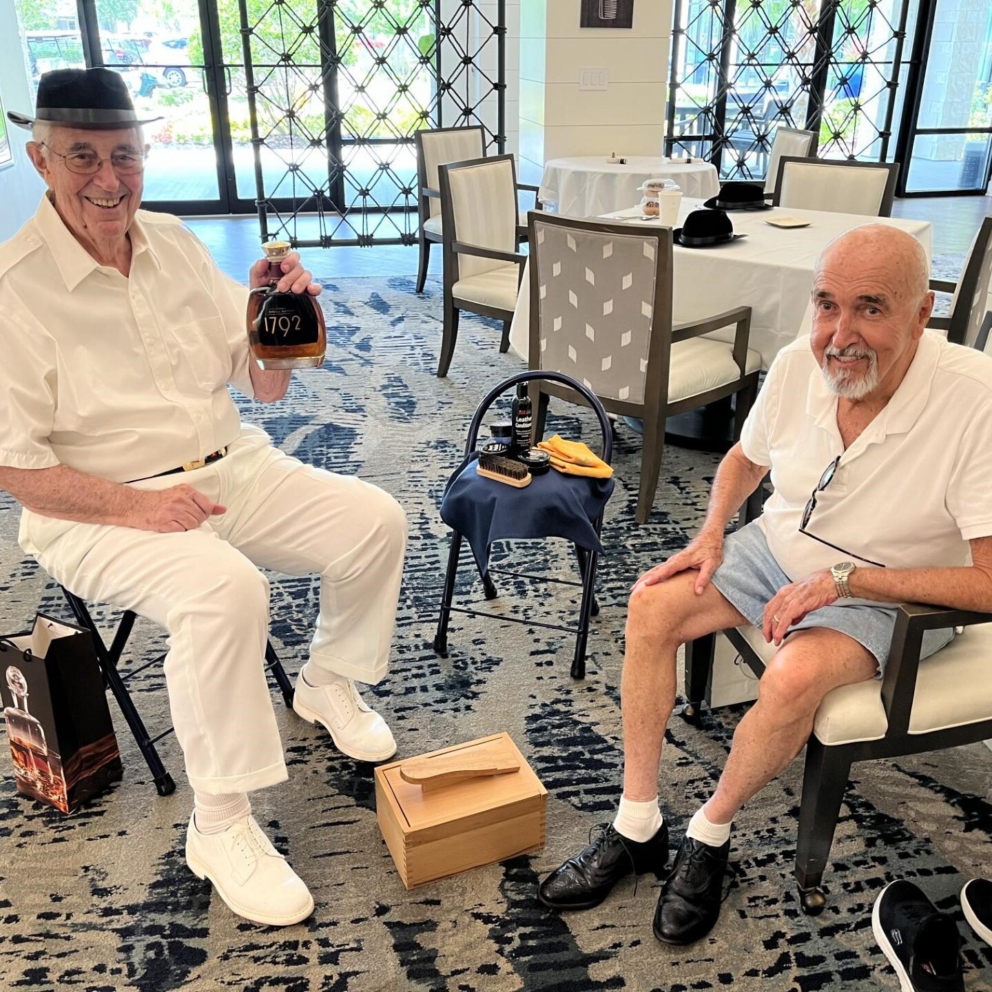 Two residents relax in a bright lounge area, one holding a bottle of bourbon, beside a shoeshine kit and neatly set dining tables at a senior living community in Athens, GA.