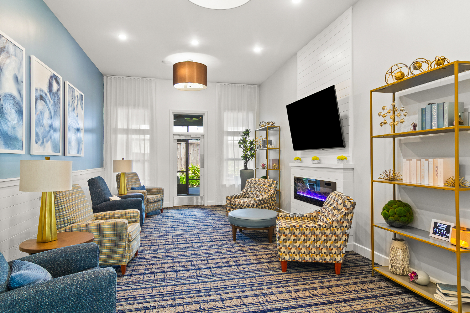A welcoming living room space with a mounted TV, gold shelving, patterned rug, and blue accent chair offers a cozy and stylish retreat at The Lakeside at Amelia Island.