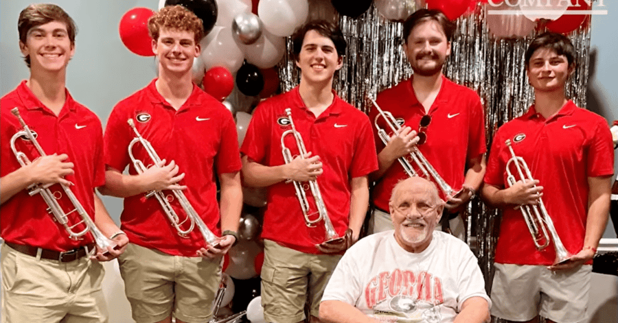Five young men in red University of Georgia polo shirts and khaki shorts stand holding silver trumpets behind a smiling older man seated in a chair, wearing a white Georgia T-shirt and a nasal oxygen cannula, in front of red, black, white, and silver balloons and a silver tinsel backdrop.