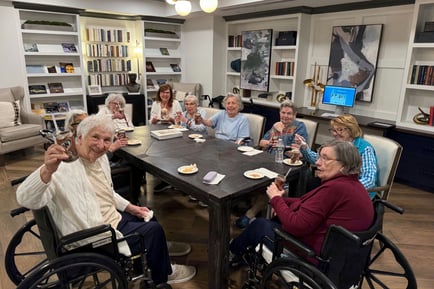 A cheerful group of older adults raises their glasses together around a large table in a book-filled common room, enjoying a lively social gathering at a senior living community in Athens, GA.