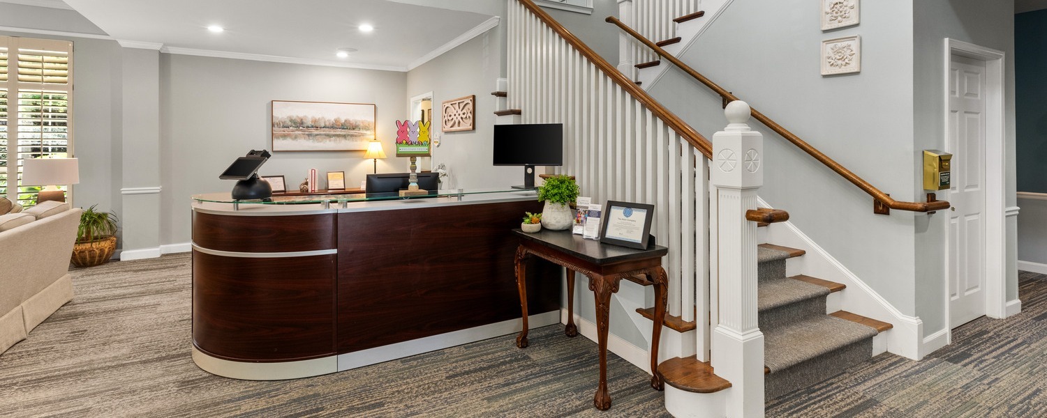 A welcoming reception desk with a curved wood counter and staircase at Arbor Terrace Athens, a senior living community in Athens, GA, reflecting resort-style senior living in Athens, GA.