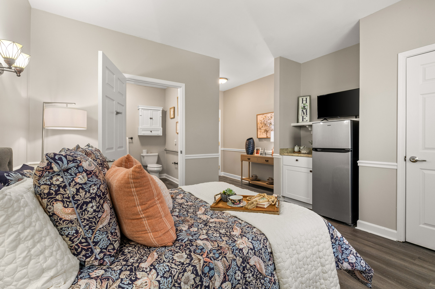 A comfortable model apartment bedroom at Arbor Terrace Athens with a cozy patterned bedspread, warm wood floors, and a glimpse of a kitchenette, showcasing everyday living at this senior community in Athens, GA.