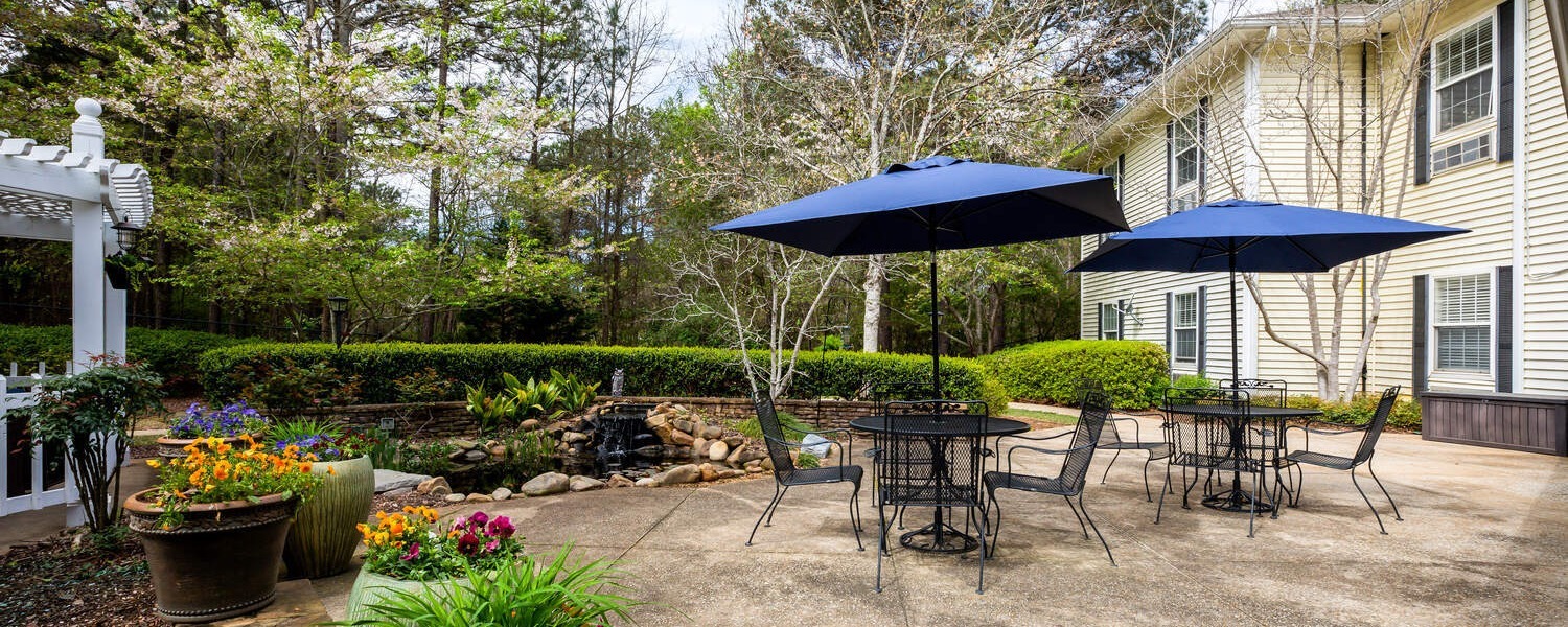 A serene courtyard patio at Arbor Terrace Athens features shaded dining tables, colorful garden beds, and a koi pond surrounded by mature trees, perfect for outdoor senior living in Athens, GA.