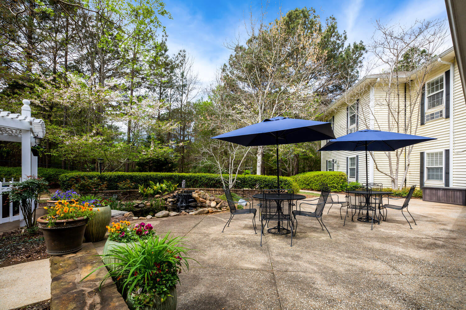 A sunny outdoor seating area at Arbor Terrace Athens with a shaded patio table, colorful flower beds, and mature trees, providing a relaxing retreat for residents of this senior living community in Athens, GA.