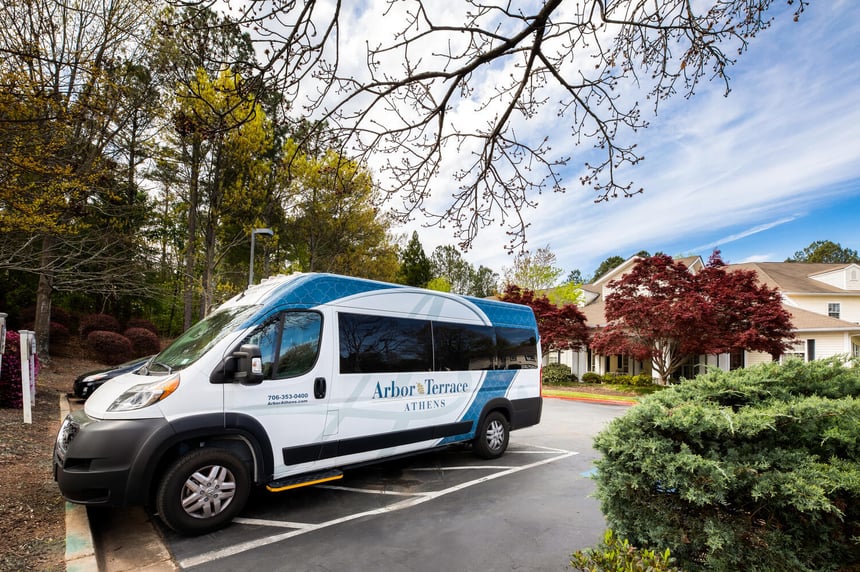 The Arbor Terrace Athens community shuttle is parked outside the senior living community in Athens, GA, surrounded by colorful trees and mature landscaping on a bright spring day.