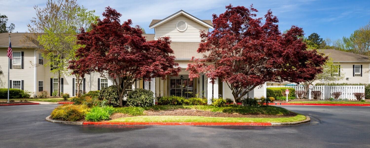 The beautifully landscaped exterior of Arbor Terrace Athens, featuring vibrant red maple trees, colorful flower beds, and a welcoming circular drive — a charming retirement community in Athens, GA.