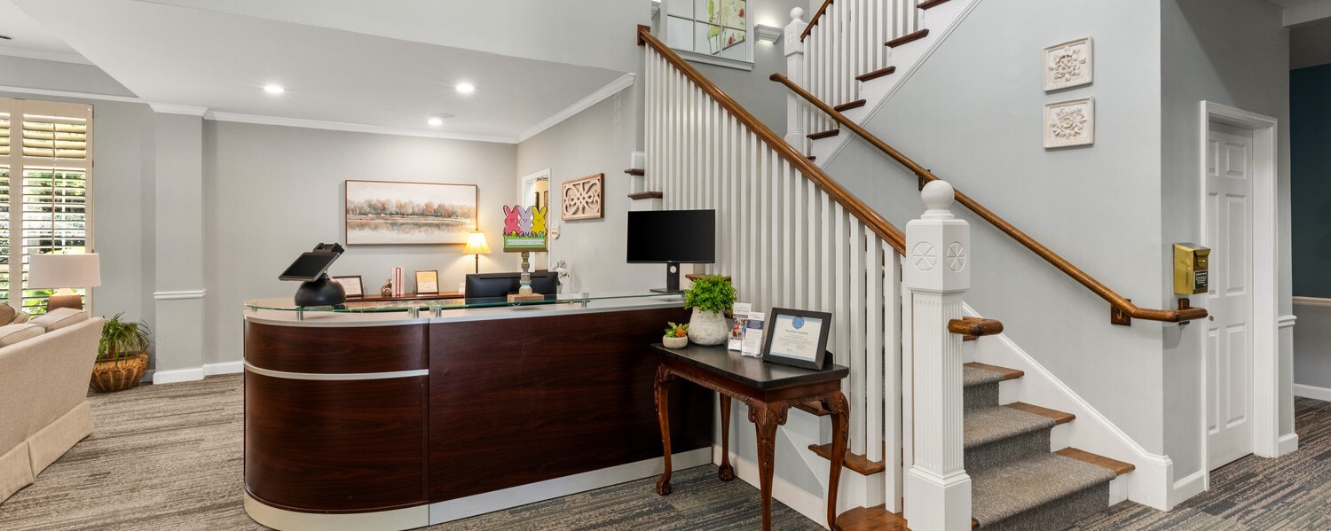 A welcoming lobby at Arbor Terrace Athens featuring a warm wood reception desk, staircase, and natural light — a gracious entry point to one of the premier senior living communities in Athens, GA.