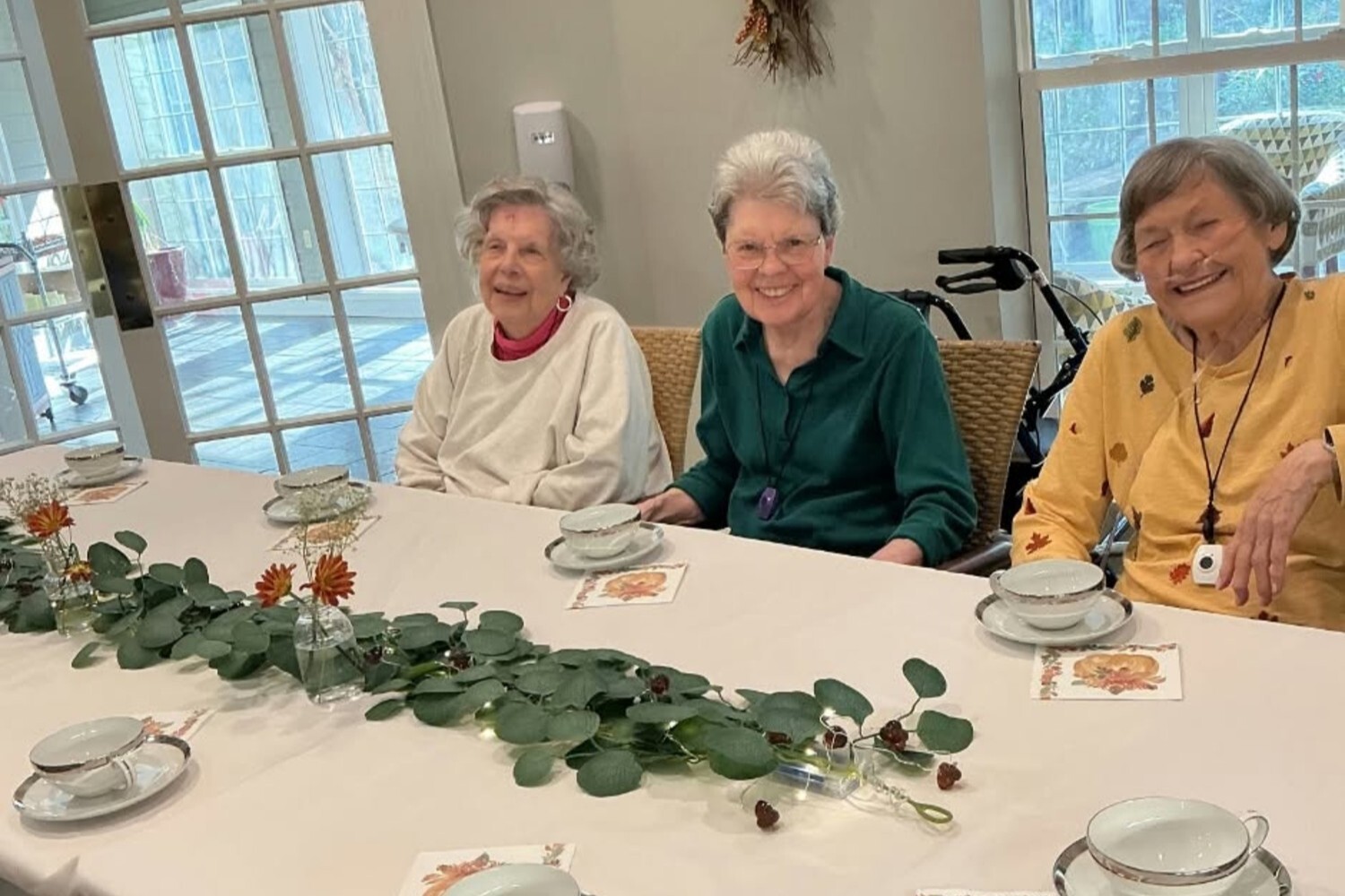 Three residents smile at a table set for a fall tea with a floral runner and teacups at Arbor Terrace Athens, a senior living community in Athens, GA, reflecting social engagement in Athens, GA.