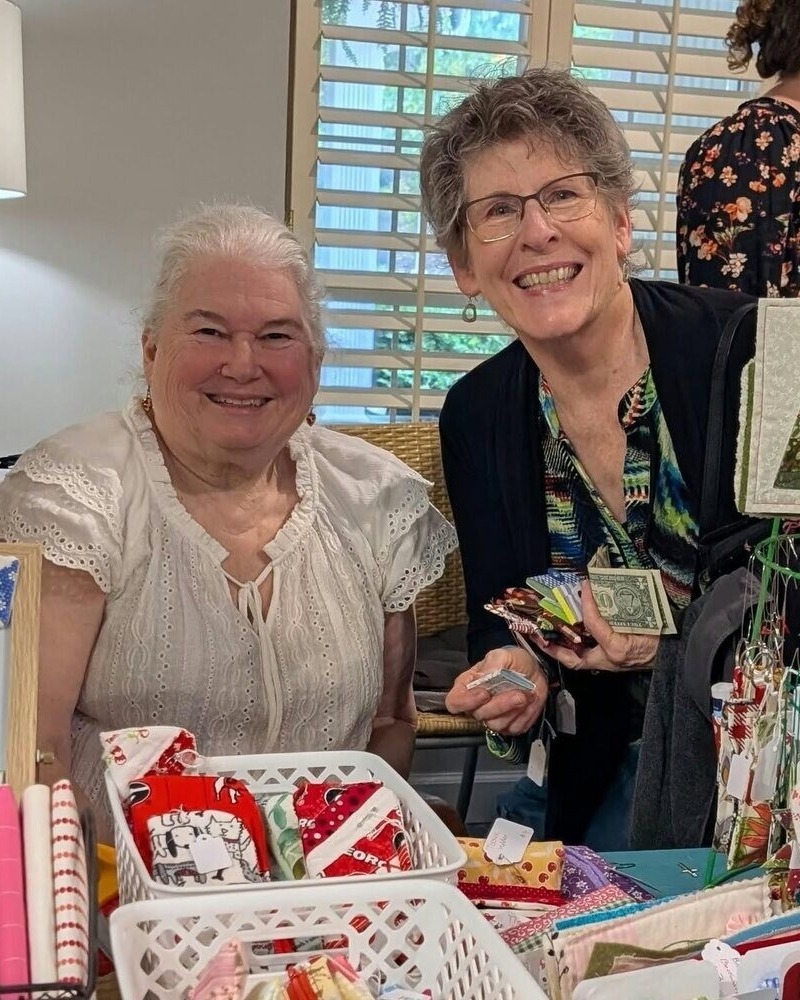 Two women smile together at a craft vendor table filled with handmade fabric items and gift bags at a senior living community in Athens, GA.