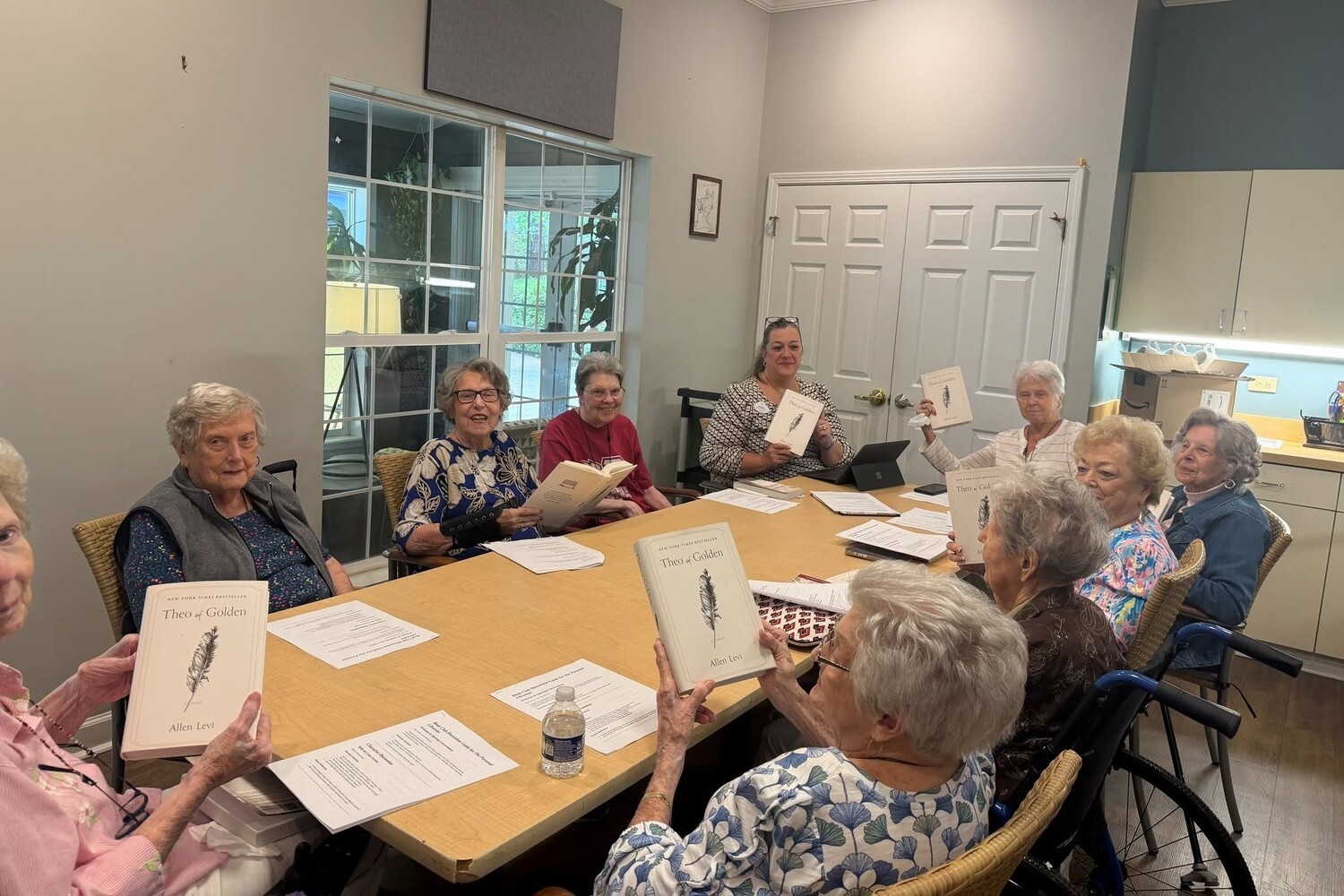 A lively book club group holds up their copies of the same novel around a large table at Arbor Terrace Athens, enjoying social engagement at a senior living community in Athens, GA.