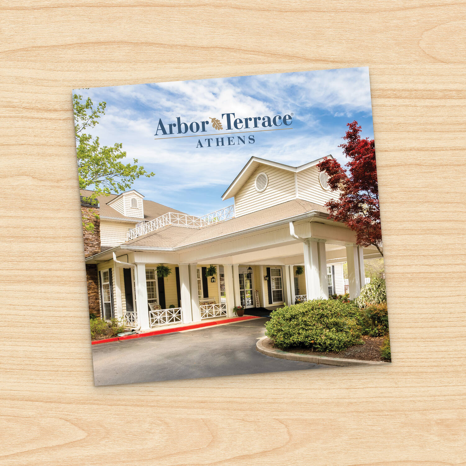 Square brochure cover for Arbor Terrace Athens senior living community, resting at an angle on a light wood surface. The cover photo shows the community's cream-colored building with a white-columned covered entrance, a red curb, and landscaped shrubs and trees under a blue sky with scattered clouds