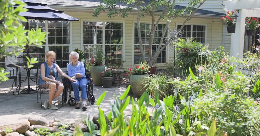 Two women enjoy a sunny afternoon together in a lush garden courtyard at a senior living community in Athens, GA, surrounded by colorful blooms and mature greenery.