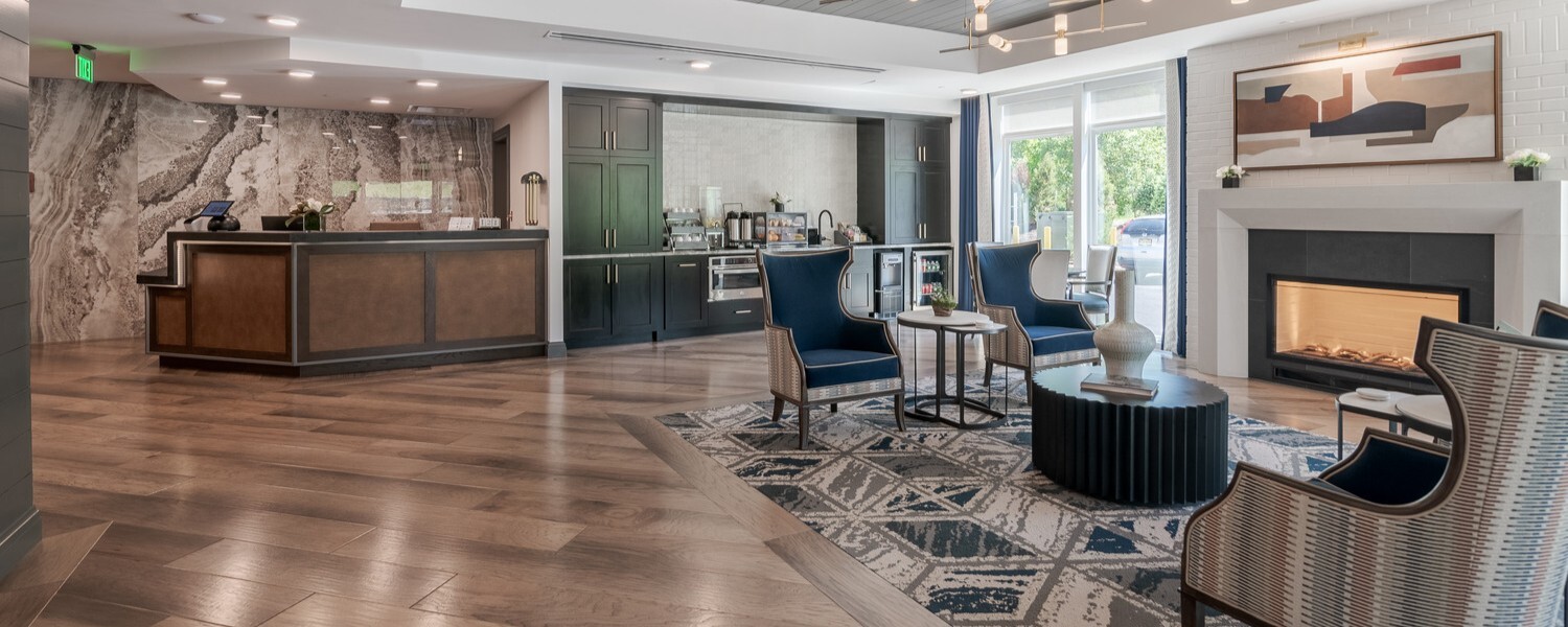 Welcoming lobby at Arbor Terrace Basking Ridge, one of the assisted living communities in Basking Ridge, NJ, with a stone-accented reception desk, plank floors, a beverage counter, and a cozy fireplace lounge with navy armchairs and a patterned rug for senior living in Basking Ridge, NJ.