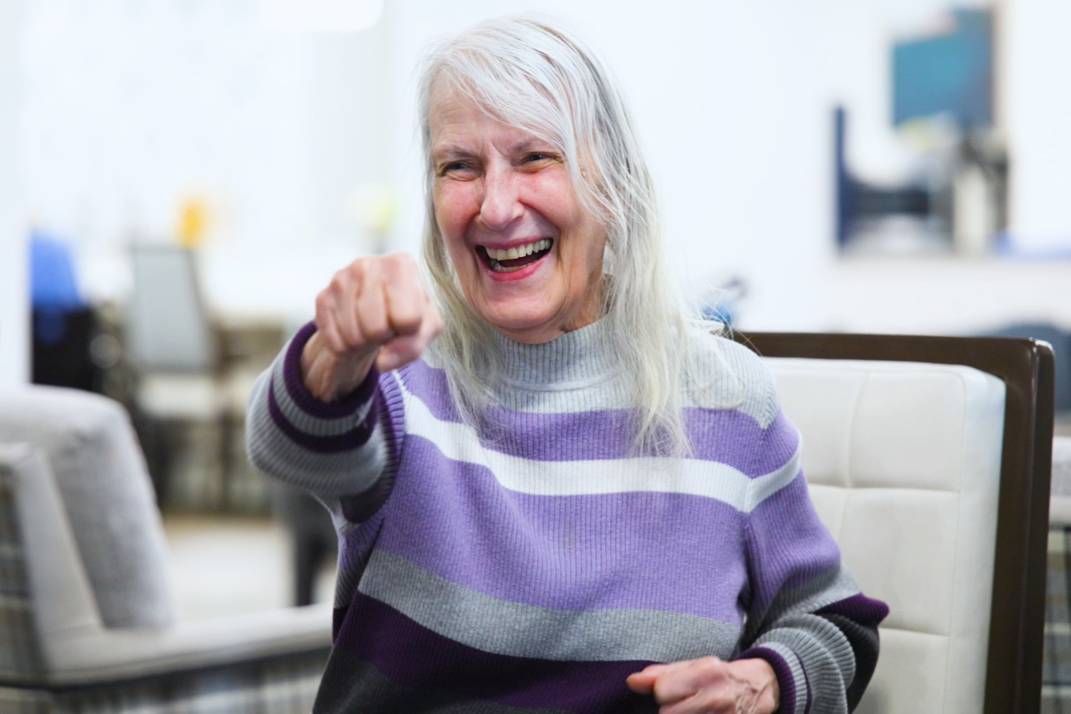 A woman with long silver hair and a purple, white, and gray striped sweater laughs and playfully points her fist toward the camera, sharing a joyful moment that captures the spirit of senior living in Basking Ridge, NJ, and top-rated senior living in Somerset County, NJ.