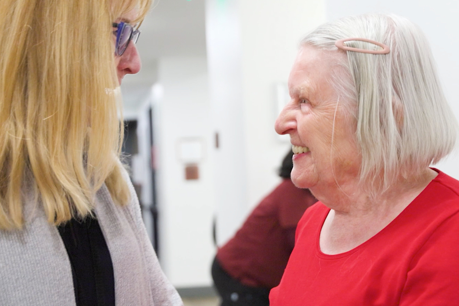A woman with short white hair and a red top smiles warmly while chatting with a blonde woman wearing glasses, a friendly moment of connection for senior living near Bedminster, NJ, and senior living near Liberty Corner, NJ.