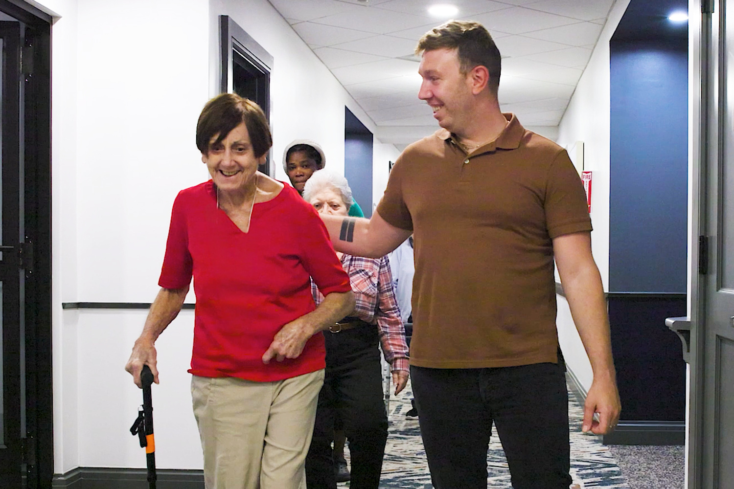 A smiling woman with short brown hair in a red top and khaki pants strolls down a bright hallway beside a smiling man in a brown polo as others follow behind, a friendly walk together at senior living in Basking Ridge, NJ, and senior living near Mendham, NJ.