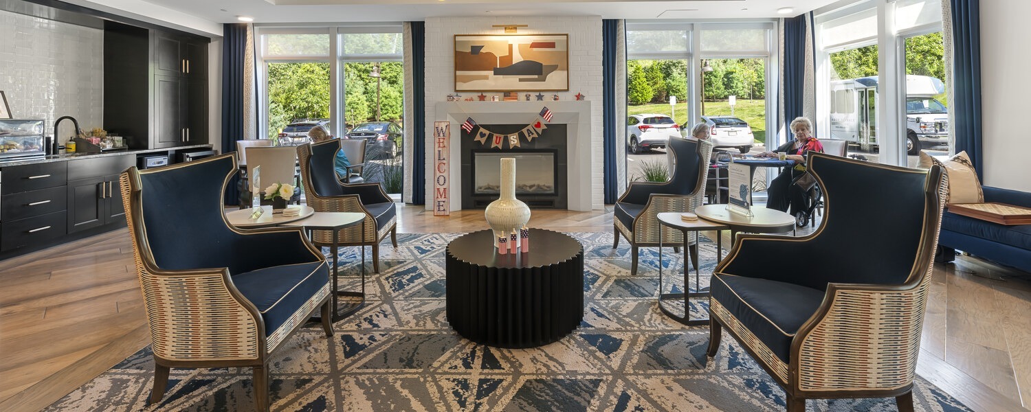 A sunlit fireplace lounge with navy wingback chairs, woven accents, a black drum coffee table with a white vase, a patterned rug, and large windows offers a cozy place to gather at senior living in Basking Ridge, NJ, and senior living near Liberty Corner, NJ.