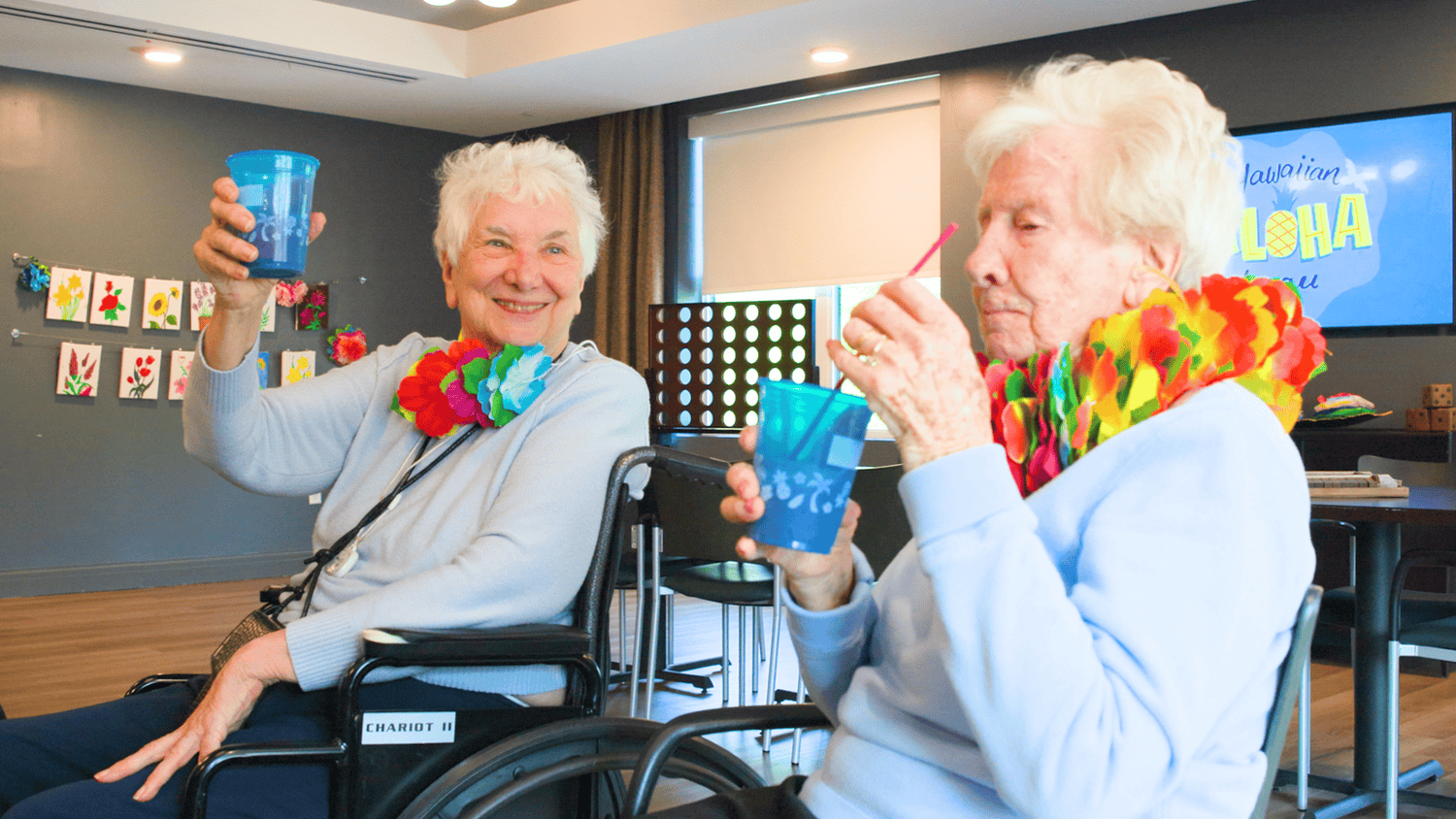 Two women with white hair and colorful leis raise blue cups in a cheerful toast at a festive gathering, celebrating resort-style senior living Basking Ridge, NJ, and senior living near Mendham, NJ.