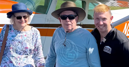 Three people posing together outdoors in front of a small orange and white propeller plane. On the left, an older woman with white hair wearing a blue sun hat, sunglasses, and a floral long-sleeve shirt holds hands with an older man in the center. The man wears a wide-brimmed tan hat, sunglasses, and a light blue 