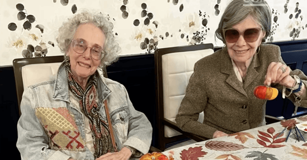 Two smiling senior women sit side by side at a table covered in an autumn leaf print. The woman on the left has curly white hair and wears glasses, a patterned scarf and a denim jacket with colorful embroidered patches. The woman on the right wears tinted sunglasses and a brown tweed blazer, and she holds up a small pumpkin painted orange and red. Paintbrushes and another painted pumpkin are on the table, and the wall behind them has a floral wallpaper print.