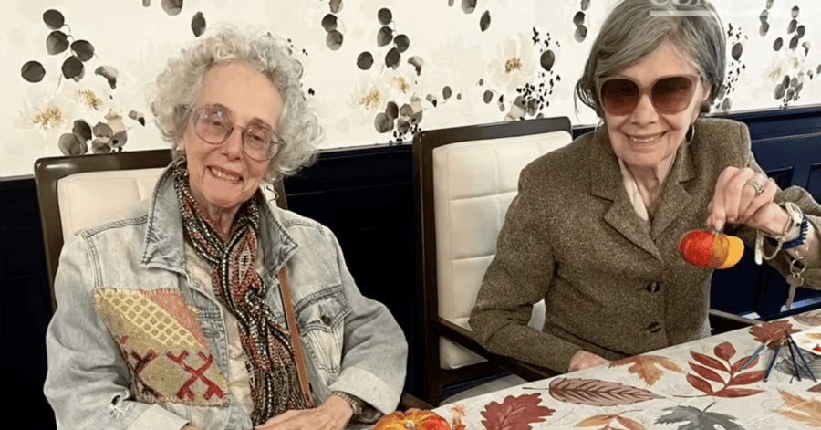 Two smiling senior women sit side by side at a table covered in an autumn leaf print. The woman on the left has curly white hair and wears glasses, a patterned scarf and a denim jacket with colorful embroidered patches. The woman on the right wears tinted sunglasses and a brown tweed blazer, and she holds up a small pumpkin painted orange and red. Paintbrushes and another painted pumpkin are on the table, and the wall behind them has a floral wallpaper print.