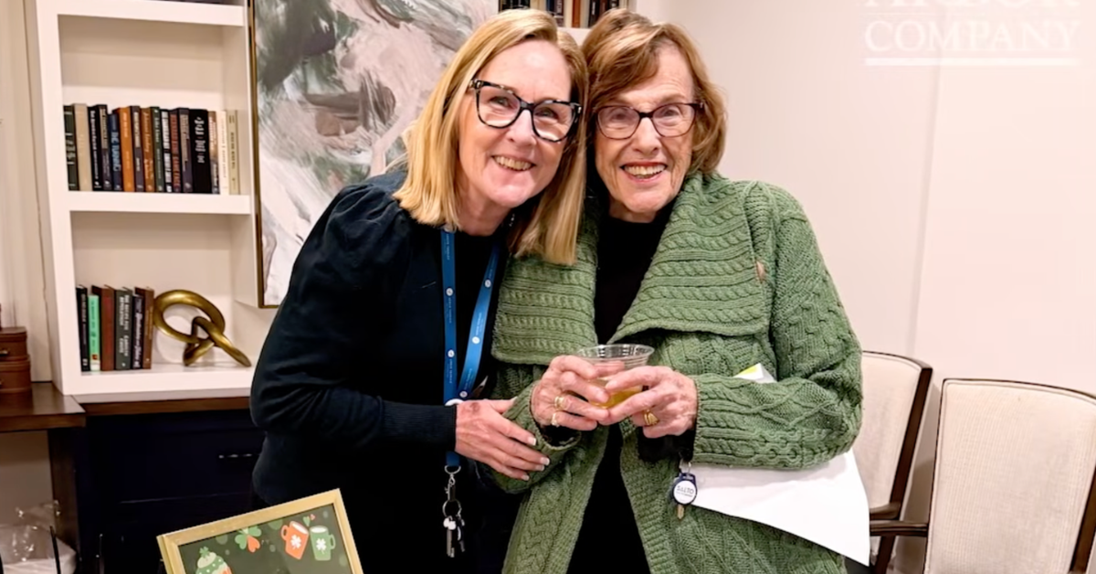 Two women pose together and smile at the camera at what appears to be a social or networking event. The woman on the left has shoulder-length blonde hair, wears dark-framed glasses, a black top, and a blue lanyard. The woman on the right is older, with short reddish-brown hair, dark-framed glasses, a black top, and a green cable-knit cardigan; she holds a clear plastic cup with a drink. They stand in front of a bookcase filled with books and a framed landscape painting.