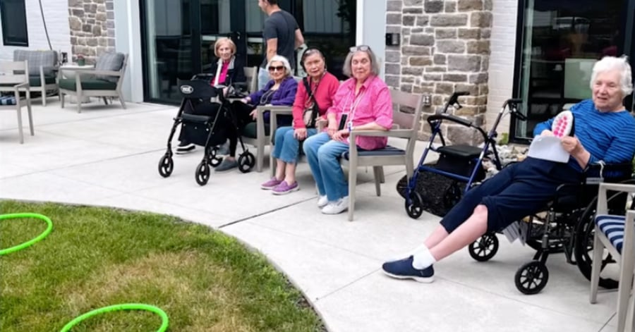 Four seated elderly women, three using rollator walkers, smile at the camera on a patio, while another woman in a wheelchair holds up a card in the foreground; green ring-toss hoops lie on the lawn nearby.