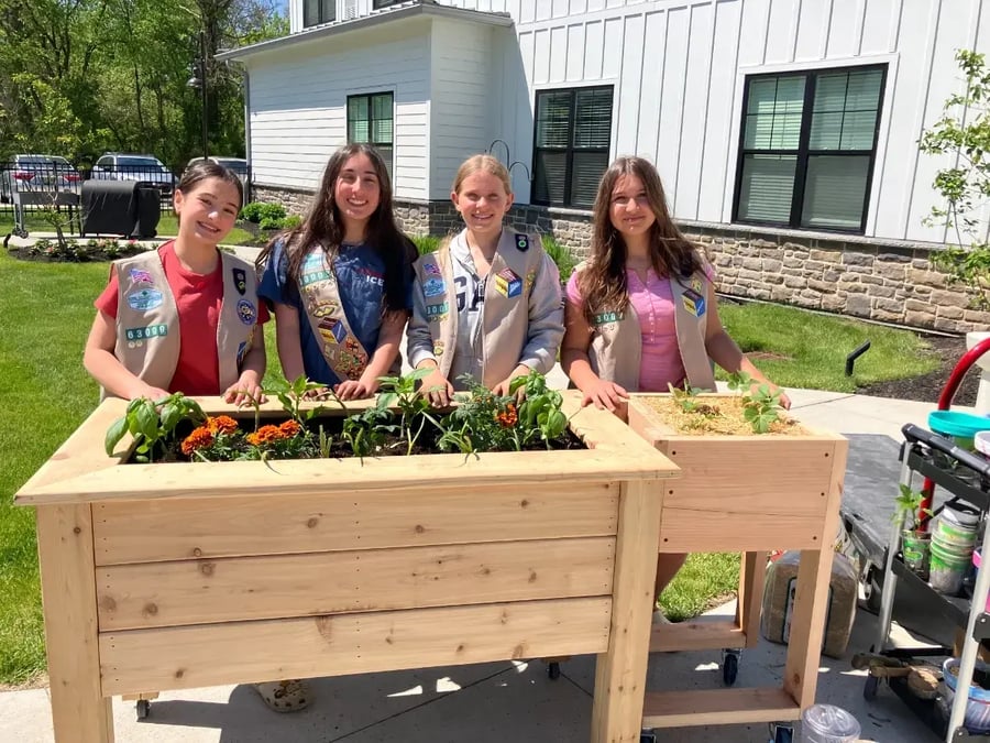 Four Girl Scouts wearing tan vests with merit badges stand behind two raised wooden garden beds on a sunny day outside a white building. The larger planter contains blooming marigolds, basil, and other vegetables, while the smaller planter holds young strawberry plants. Gardening tools and supplies are visible nearby, highlighting a community gardening project.