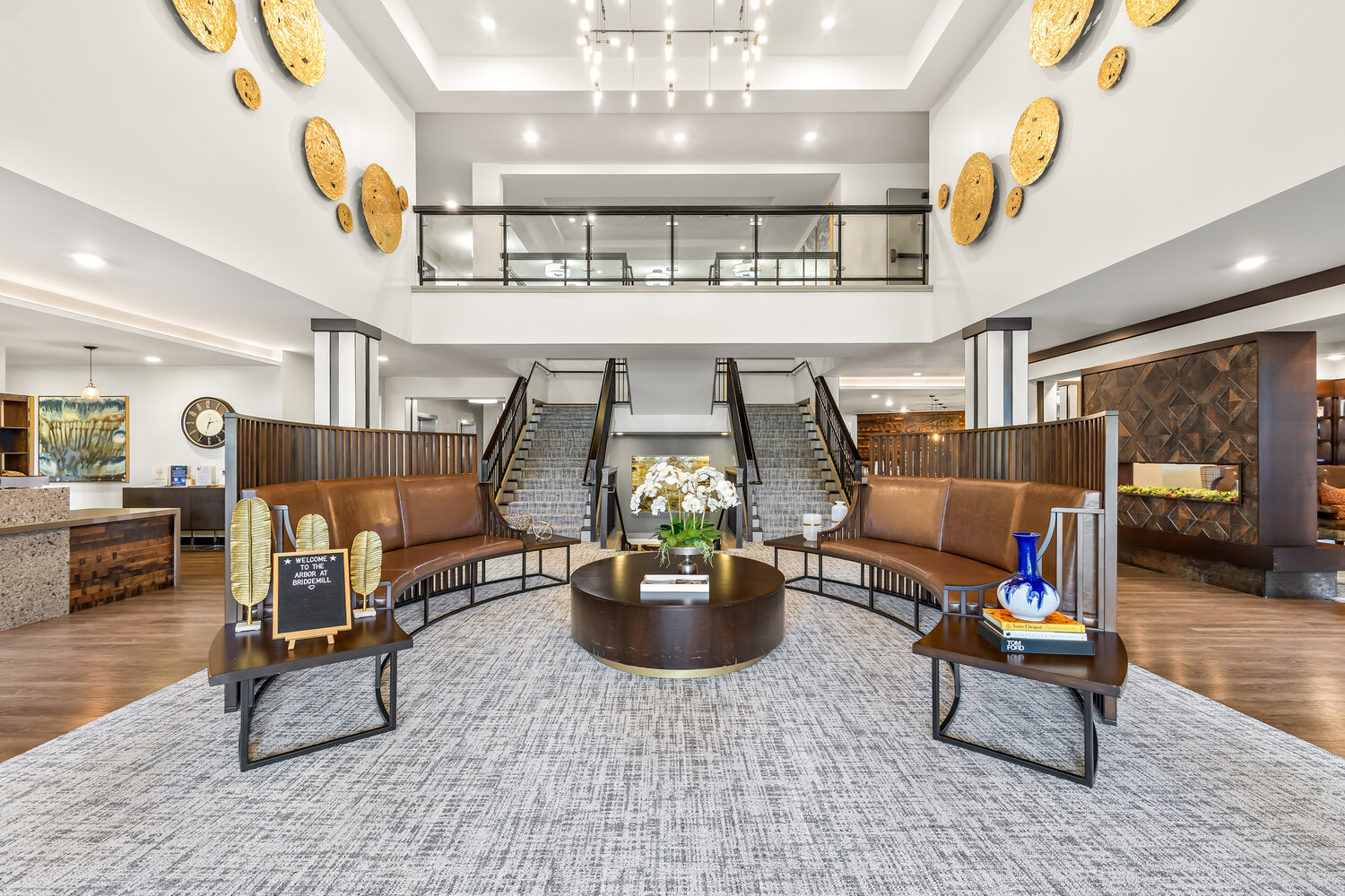 A grand two-story atrium with a sweeping staircase, decorative gold wall accents, and elegant seating at The Arbor at BridgeMill, a senior living community in Canton, GA.