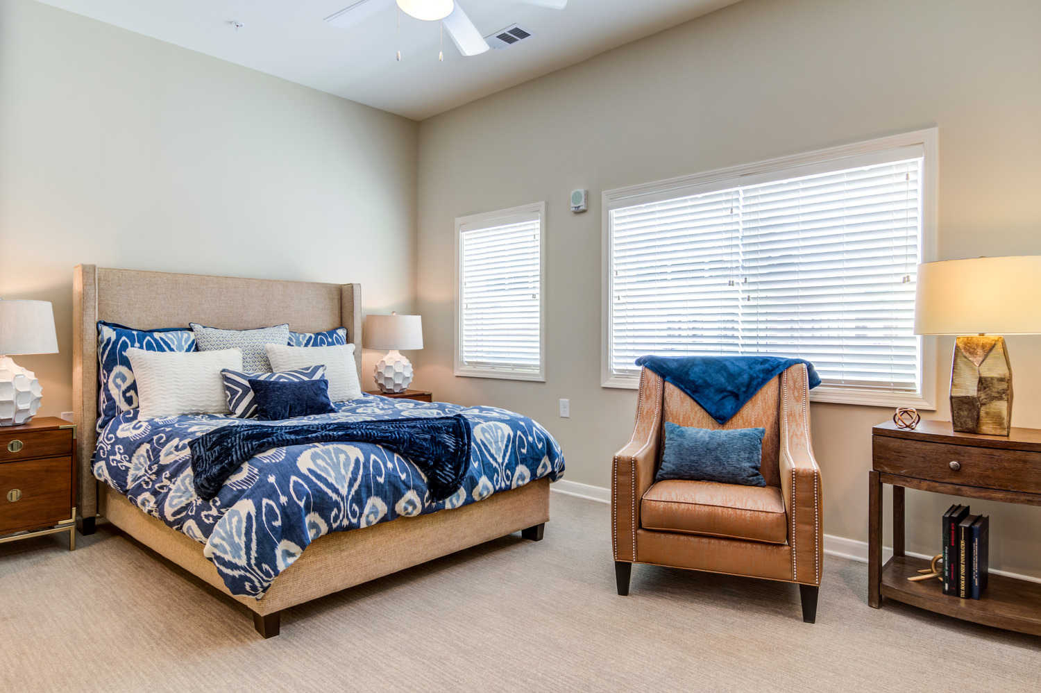 A spacious, elegantly furnished bedroom with a king bed dressed in rich blue bedding, a leather armchair, warm wood nightstands, and bright natural light in a senior apartment at Arbor Terrace Burnt Hickory in Marietta, GA.