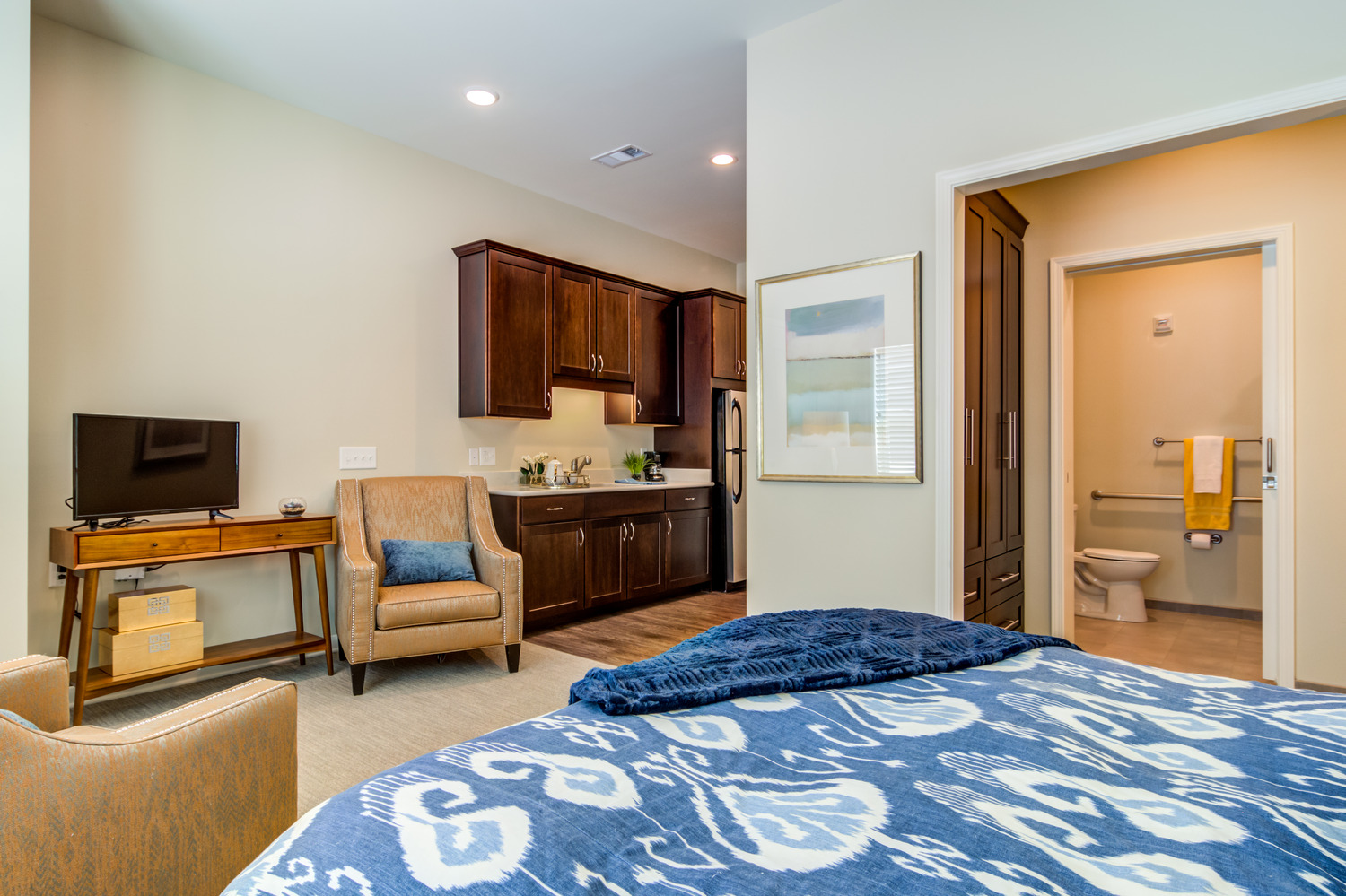 A comfortable senior apartment at Arbor Terrace Burnt Hickory in Marietta, GA, featuring a cozy sitting area, kitchenette with dark wood cabinetry, flat-screen TV, blue patterned bedding, and an accessible bathroom.