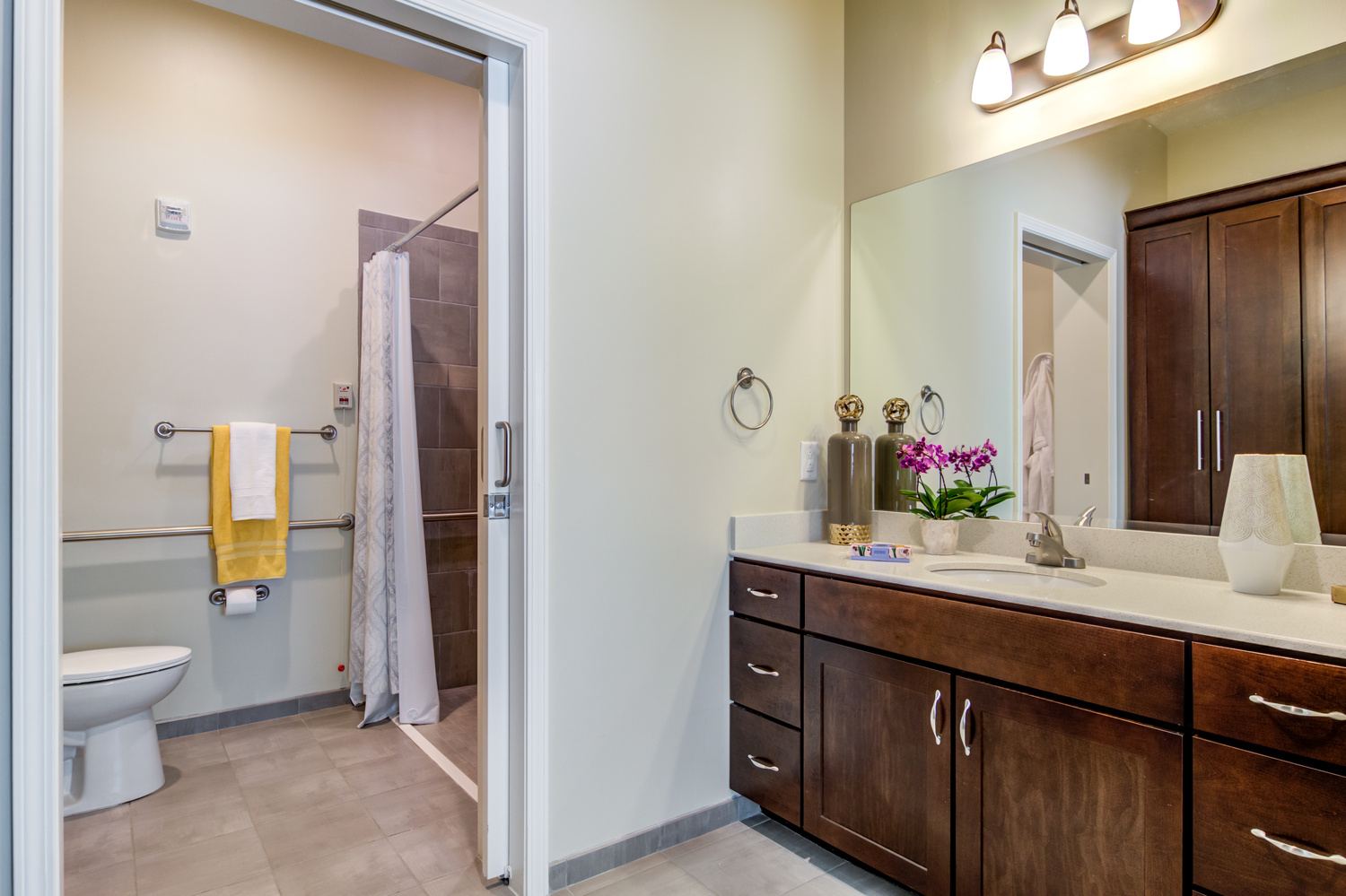 A well-designed apartment bathroom featuring dark wood cabinetry, a spacious vanity, fresh orchids, and an accessible roll-in shower with grab bars in a senior apartment at Arbor Terrace Burnt Hickory in Marietta, GA.