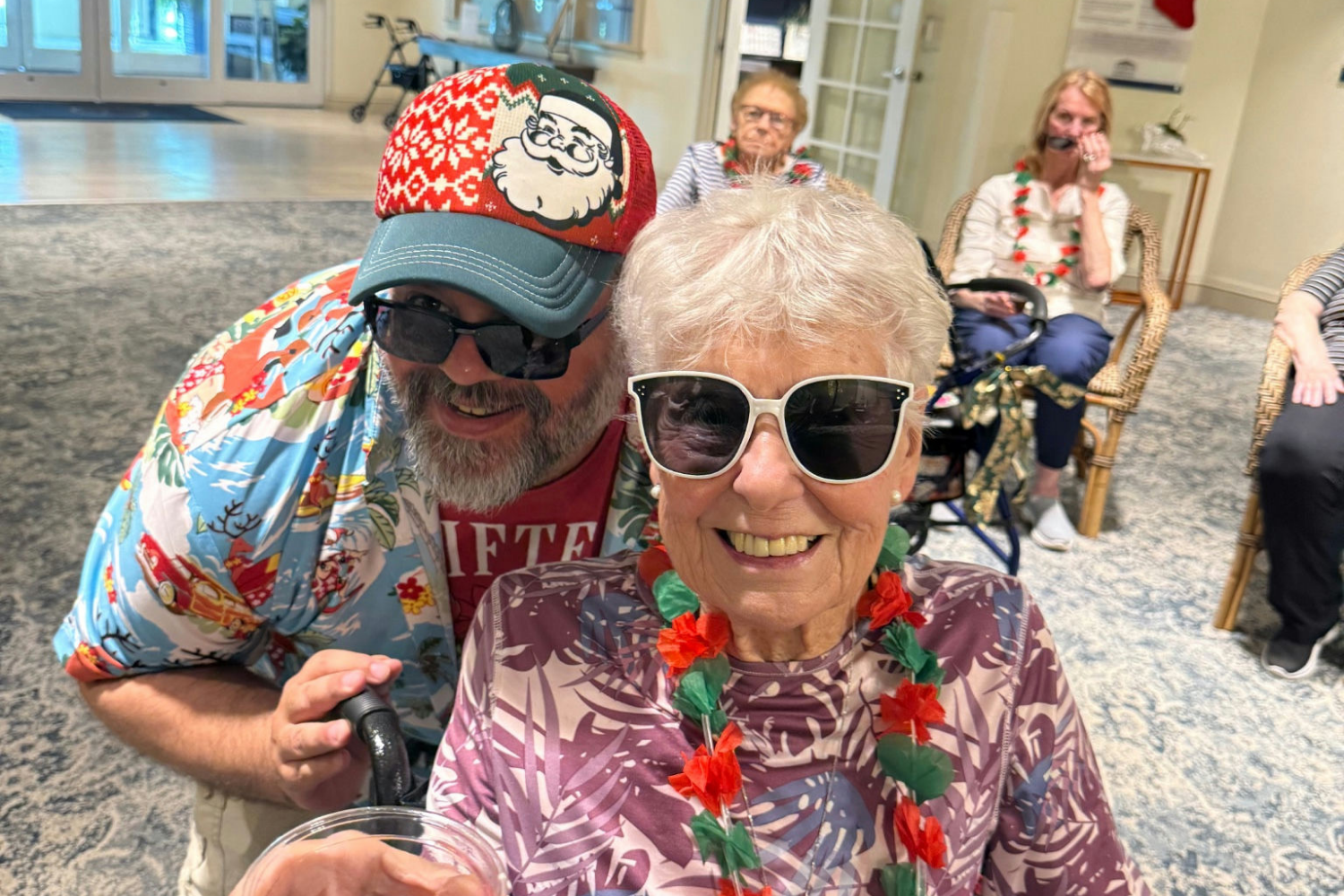 An older woman with short white hair and sunglasses smiles warmly, wearing a floral lei, seated in a wheelchair outdoors. A bearded man in a Hawaiian shirt and a red cap stands behind her, also smiling.