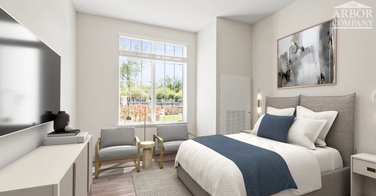 A bright, modern bedroom rendered for The Arbor Company senior living community, featuring a neatly made bed with a gray upholstered headboard, white bedding, and a navy blue accent blanket. Two gray accent chairs with gold frames sit near a large window overlooking greenery. A wall-mounted TV and white dresser are visible on the left, and an abstract framed artwork hangs above the bed on the right.