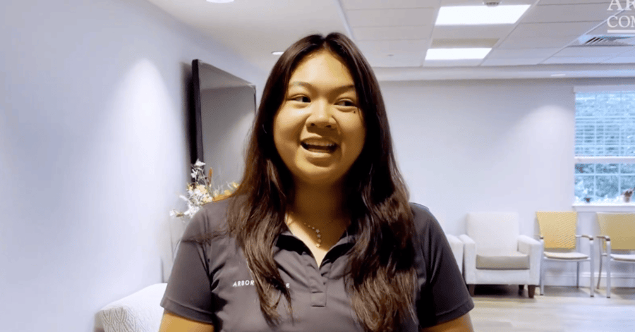A smiling team member with long dark hair, wearing a dark Arbor Terrace polo shirt, speaks to the camera in a bright, airy common area at Arbor Terrace Citrus Park, with cushioned chairs and a sunlit window behind her.