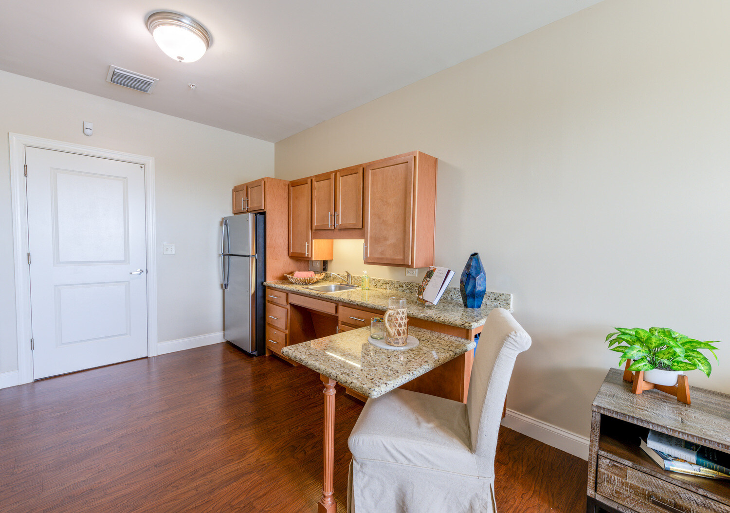 This apartment kitchenette features a granite peninsula, wood cabinetry, and a stainless steel refrigerator, showcasing the comfortable senior apartments in Cooper City, FL, at this senior living community in Cooper City, FL.