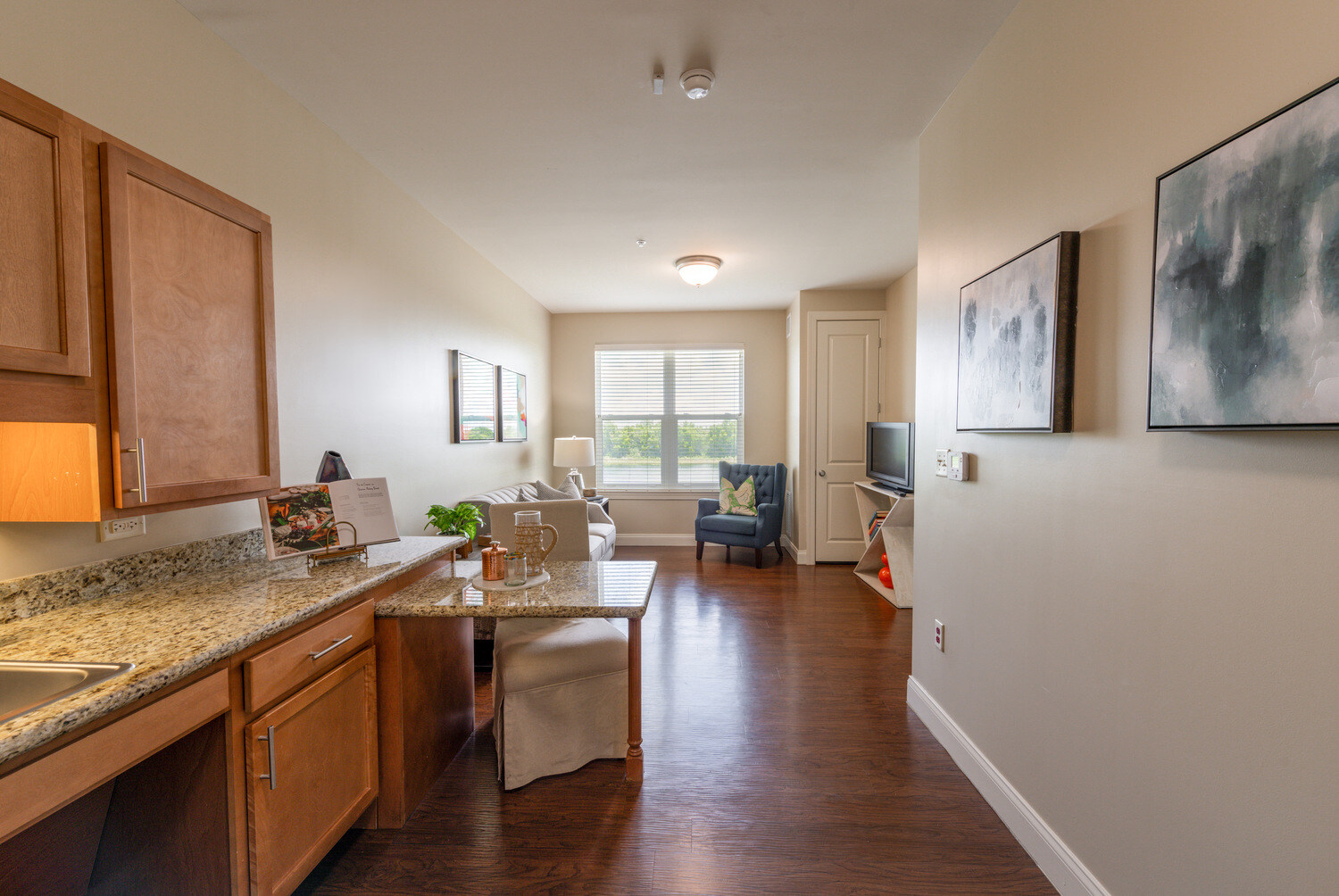 This apartment kitchenette opens into a bright living area with a blue accent chair and a TV, showcasing the comfortable senior apartments in Cooper City, FL, at this senior living community in Cooper City, FL.