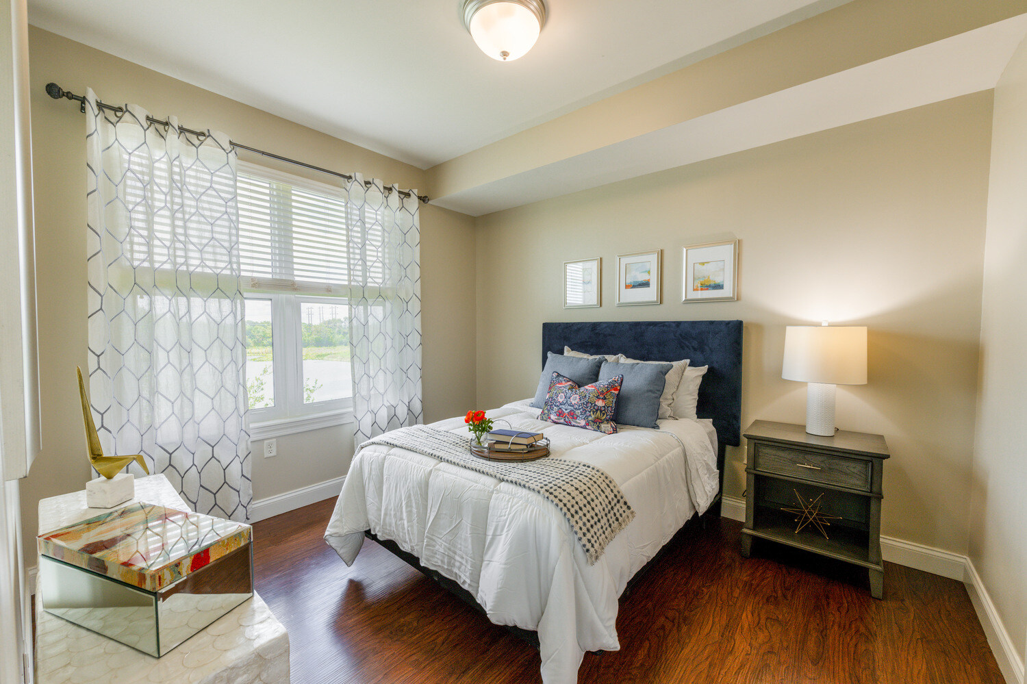 This bedroom features a plush navy upholstered headboard, sheer patterned curtains, and a bright window view, reflecting the comfortable senior apartments in Cooper City, FL, at this senior living community in Cooper City, FL.