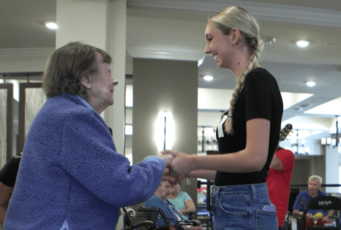A resident joyfully dances hand in hand with a smiling young woman during a lively music event, capturing the warm connections and vibrant senior living lifestyle enjoyed throughout this senior living community.