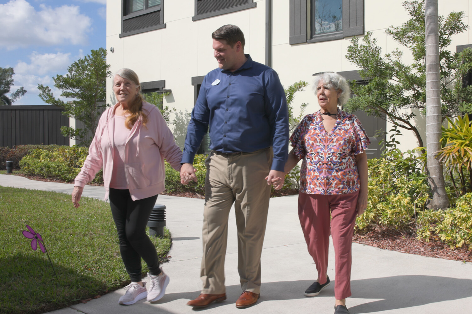 A smiling man in a navy shirt walks hand in hand with two senior women along a sunny sidewalk lined with greenery, one in a pink hoodie and the other in a colorful print top, a cheerful stroll in senior living with walkable neighborhoods Atlanta, GA and senior living in Atlanta, GA.