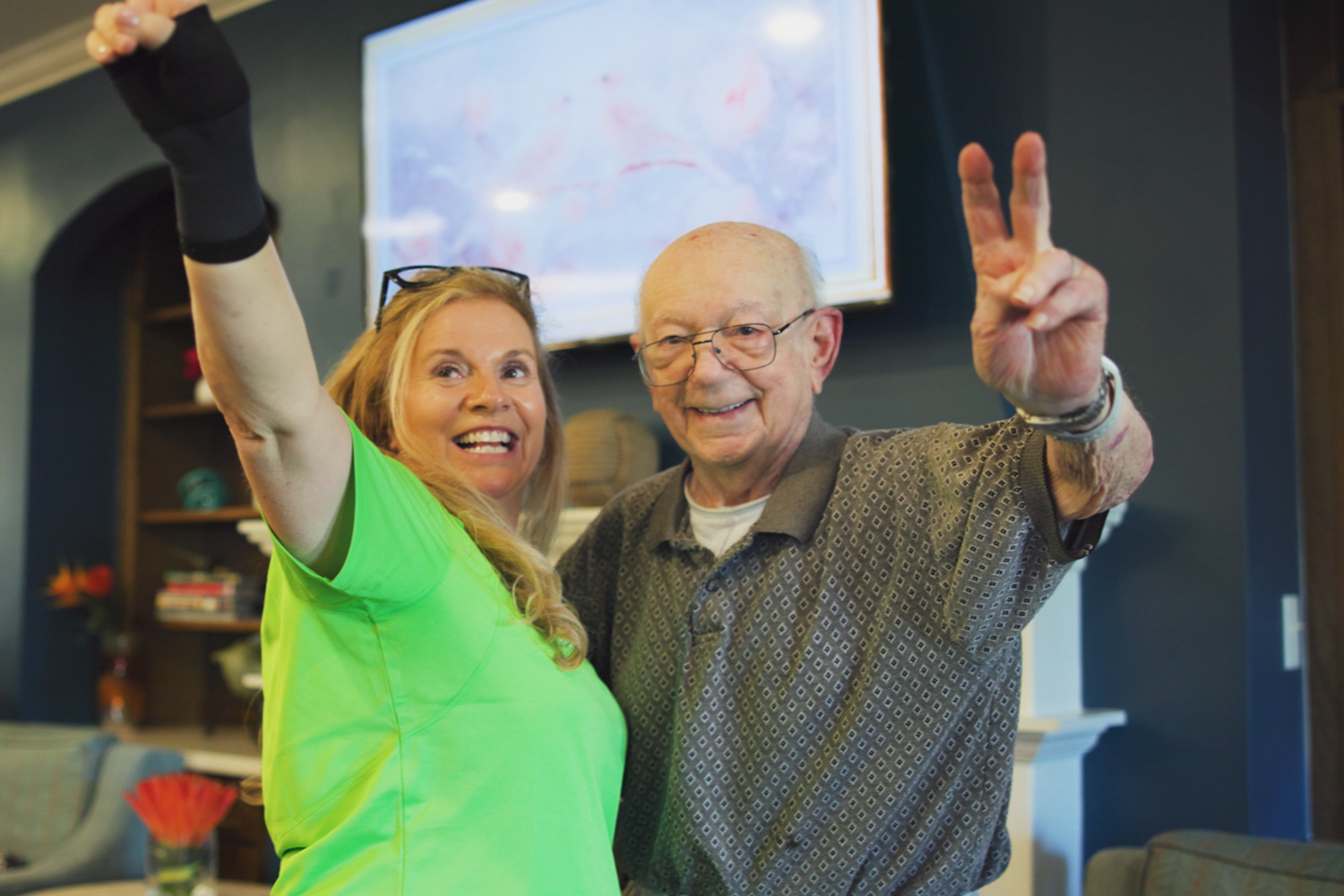 A resident throws up a peace sign while a staff member in a green top raises her arm in celebration, both smiling during a lively group activity in the lounge, reflecting senior living with social activities at senior living in Cooper City, FL.