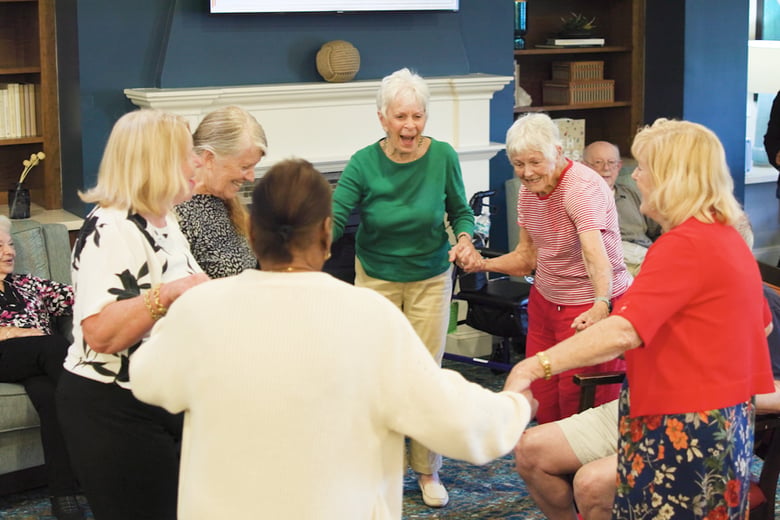 Residents dance and hold hands together indoors, sharing laughter and connection in a warm senior living community at The Arbor Company.